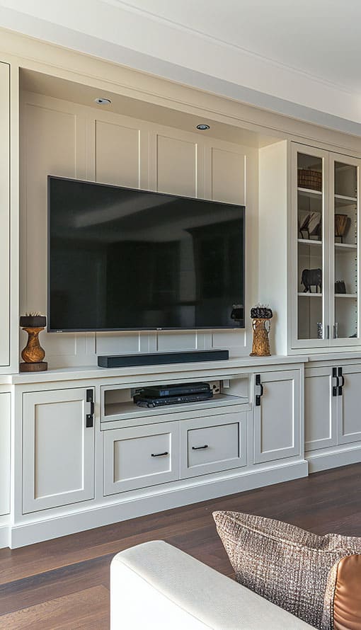 A modern living room features a large flat-screen TV mounted on a light-colored built-in wall unit. The unit has closed cabinets and open shelving with decorative items. A soundbar sits below the TV, and the room has dark hardwood floors and a partial view of a sofa.